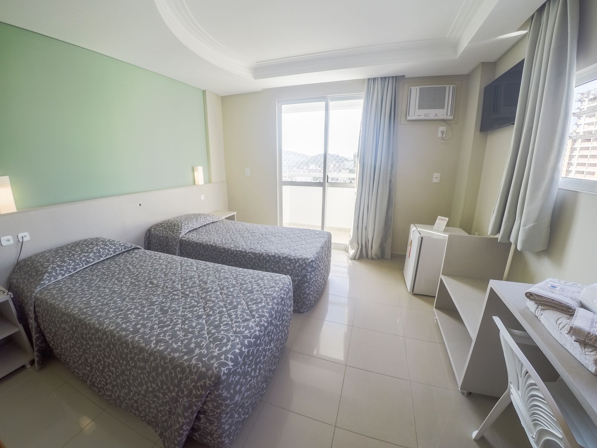 Quarto de hotel com duas camas, uma escrivaninha, uma cadeira, televisão e varanda com cortinas.