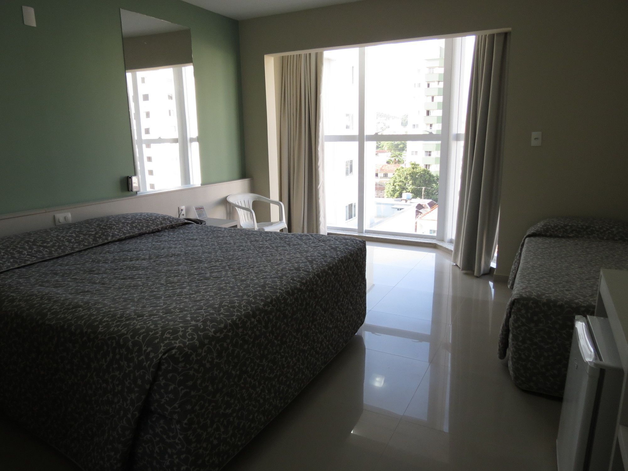 A hotel room with a large bed, a smaller bed, a desk, and a chair. There is a window with curtains and a view of buildings outside.