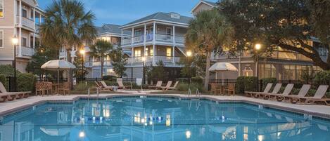 A swimming pool with lounge chairs and umbrellas, surrounded by multi-story buildings with balconies and palm trees.