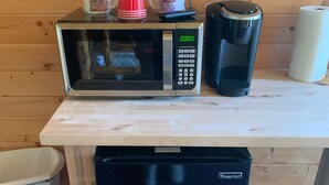 Un microondas sobre una mesa de madera con una cafetera, un cubo de basura y un refrigerador pequeño.