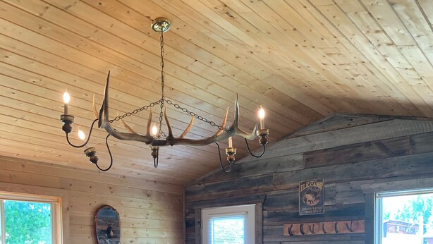 Interior rústico de una cabaña con techo de madera, un candelabro, una cama con colcha estampada, una mesa de comedor de madera y sillas.