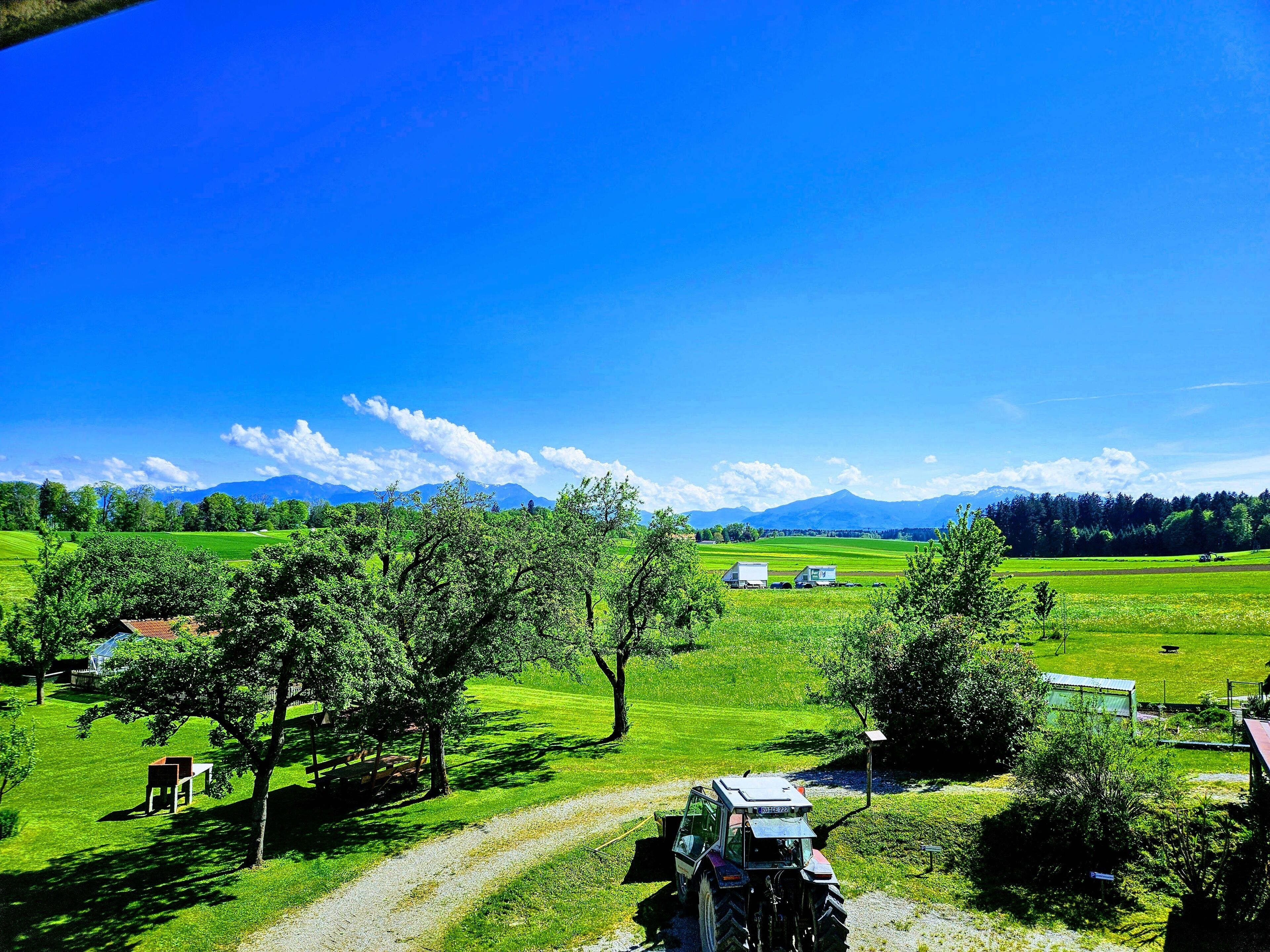 Paisaje rural con un tractor en un camino de tierra, campos verdes y montañas a lo lejos.