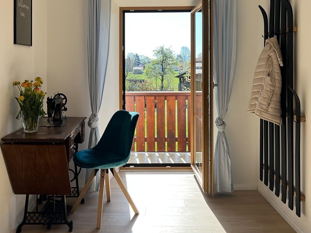 A room with a wooden desk, a blue chair, a view of a wooden deck through a glass door, and a coat rack with a jacket.