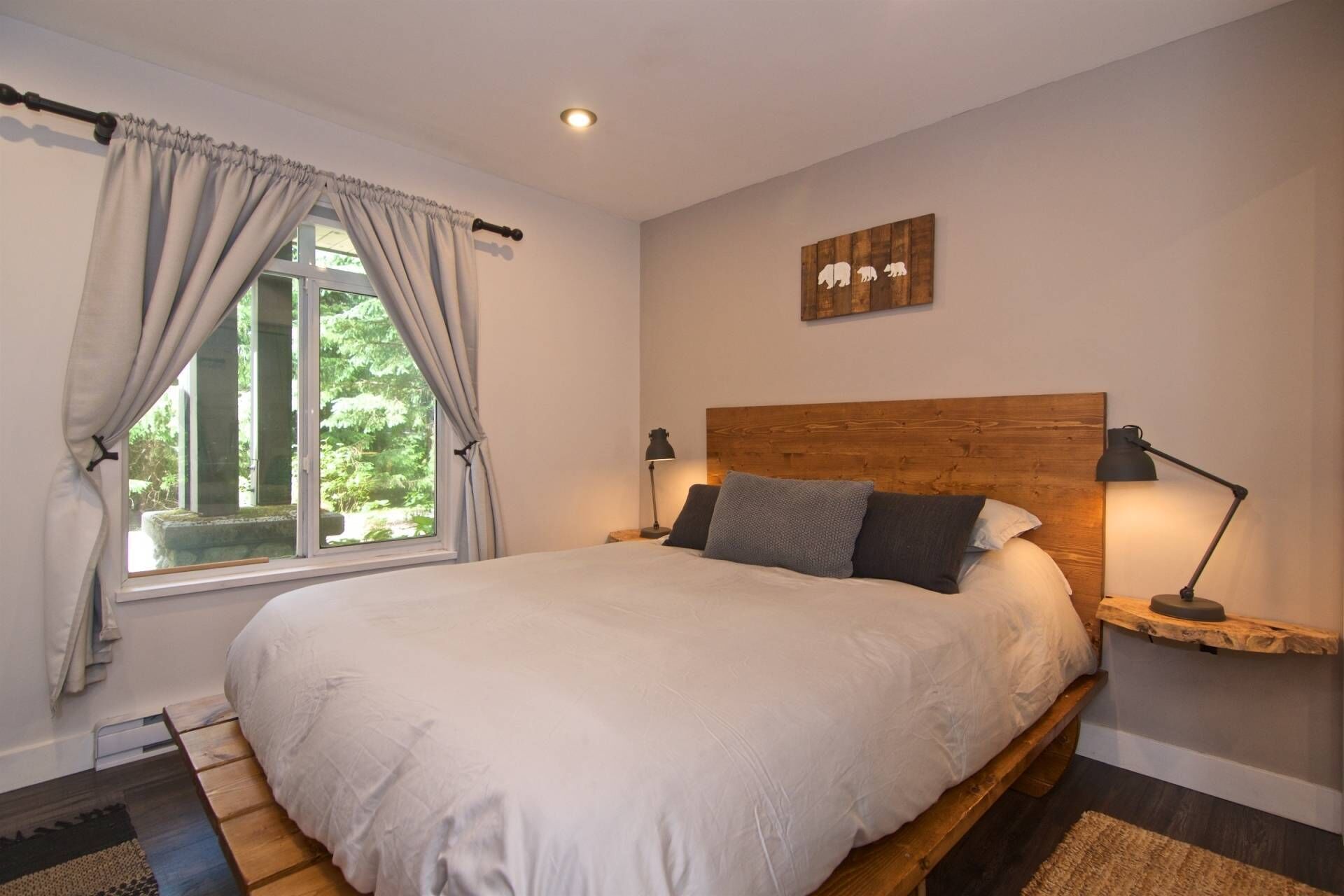 Une chambre à coucher avec un grand lit, une tête de lit en bois, une table de chevet avec une lampe, une fenêtre avec des rideaux et une œuvre d’art accrochée au mur.