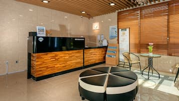 Un espace d’accueil comprenant un comptoir de réception en bois, un présentoir noir pour les informations, un coin salon rond noir et blanc, et une petite table sur laquelle est posée une plante en pot.