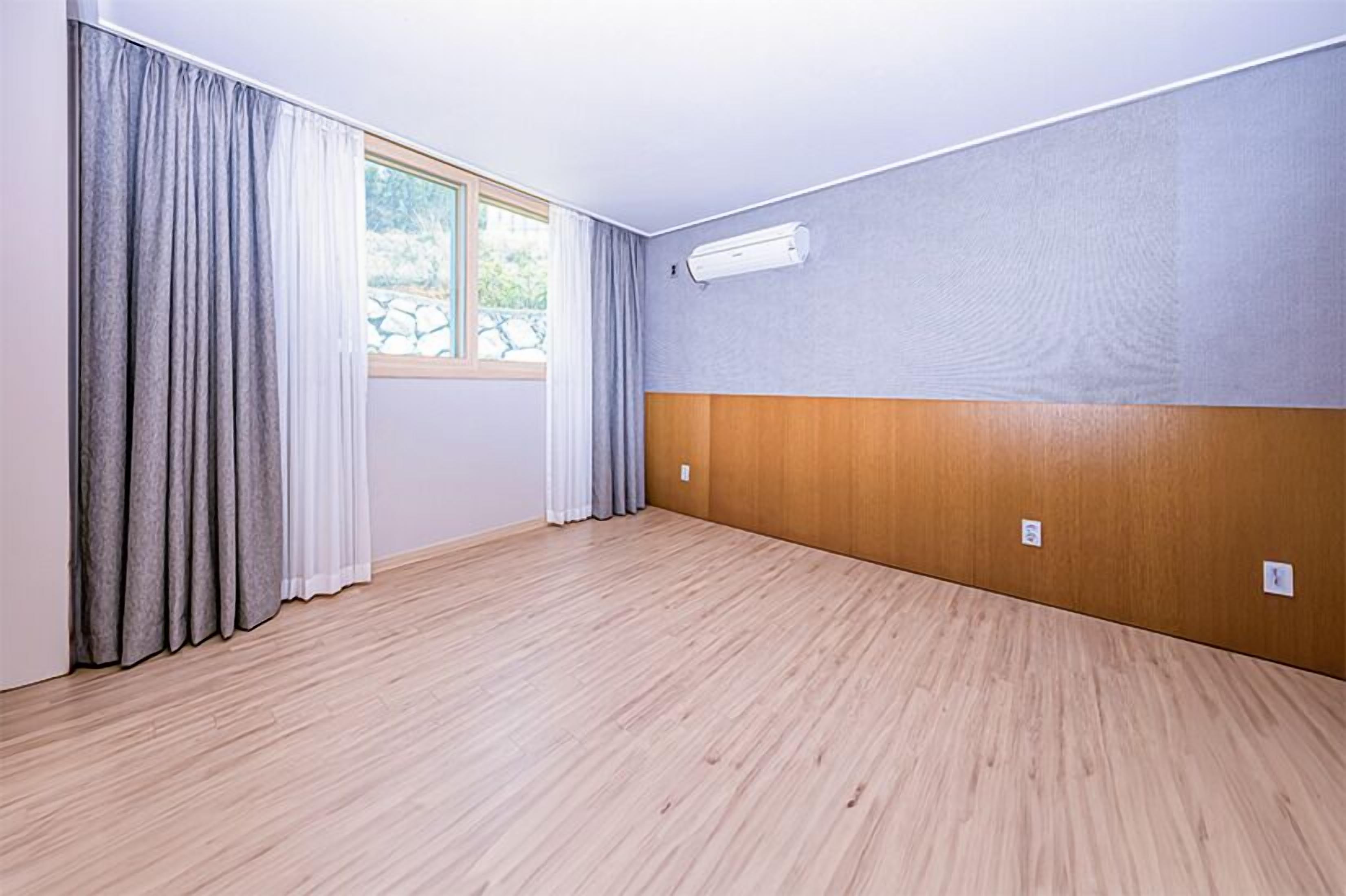Una habitación con un ventanal grande, cortinas grises, piso de madera y aire acondicionado instalado en la pared.