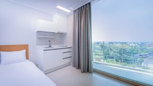 A modern hotel room with a bed, a kitchenette, and a large window offering a city view.