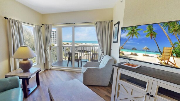 Ein Hotelzimmer mit Balkon, einem Fernseher, der eine Strandansicht zeigt, einer Couch, einem Tisch und einer Lampe.