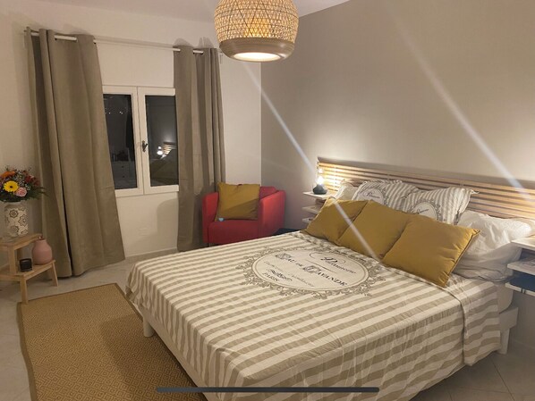 A hotel room with a bed, a nightstand, a red chair, a window with curtains, and a decorative lamp.