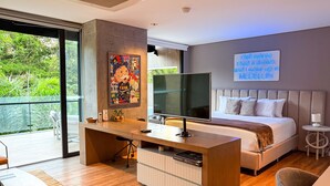 Ein modernes Schlafzimmer mit einem großen Bett, einem Schreibtisch mit Fernseher und einem Balkon mit Blick auf Grünflächen.