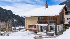 A two-story wooden house with a snow-covered roof, a visible chimney, and a glass door entrance.