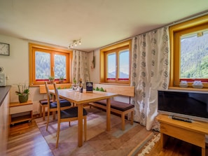 A cozy dining area with a wooden table and chairs, a TV on a wooden stand, and large windows with curtains.
