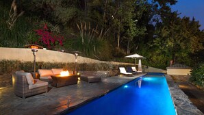 Ein Swimmingpool mit einer Steinterrasse, Liegestühlen und einer Feuerstelle.