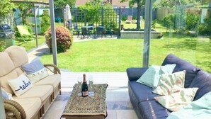 En overdækket patio med en sofa, et bord med en flaske og glas, samt udsigt til en have.