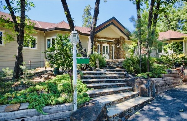 Situated in a wooded area, this two-story house features a garden with shrubs and a tree, and a stone facade with a pitched roof.