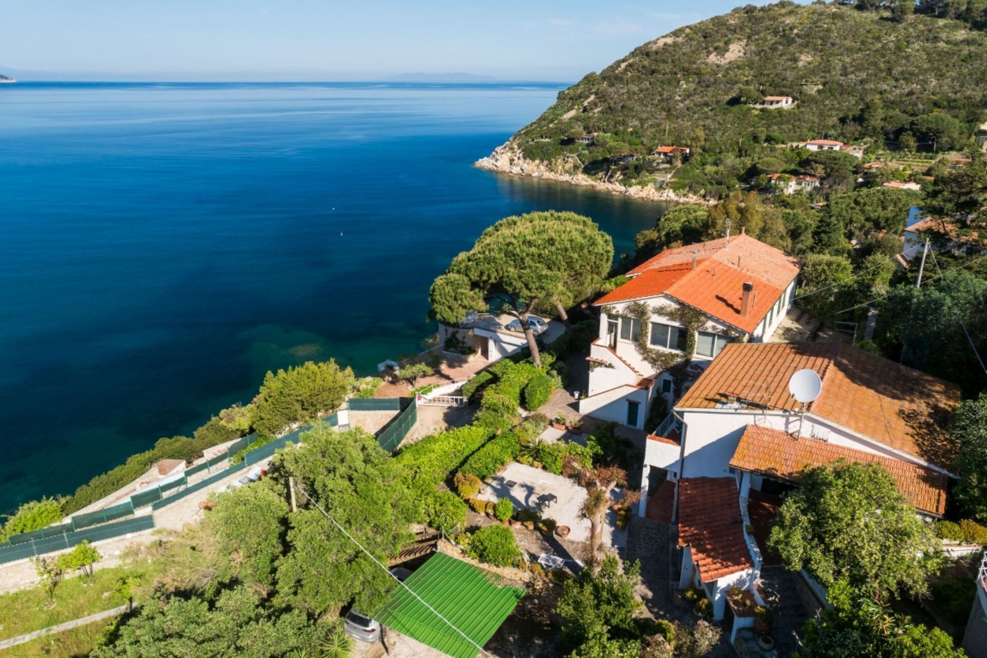 An aerial view of a coastal property with a house featuring a red-tiled roof, surrounded by lush greenery and a clear view of the sea.