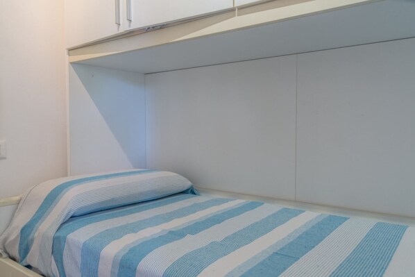 A small, single-bed room with a striped bedspread, a white wall, and a white ceiling.