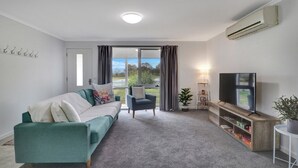 A modern living room with a teal sofa, a small armchair, a TV on a wooden stand, and a large window with curtains. - Katta Cottage (Mansfield)