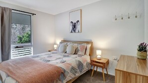 A bedroom with a bed, bedside tables, a window with curtains, a wall-mounted picture, and a small wooden desk with a plant. - Katta Cottage (Mansfield)