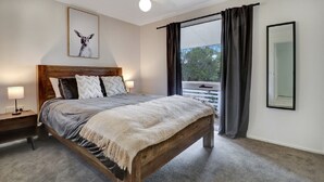 A bedroom with a wooden bed, a nightstand with a lamp, a large window with curtains, and a framed picture on the wall. - Katta Cottage (Mansfield)