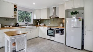 A modern kitchen with white cabinetry, a stainless steel range hood, a built-in oven, a microwave, and a refrigerator. - Katta Cottage (Mansfield)