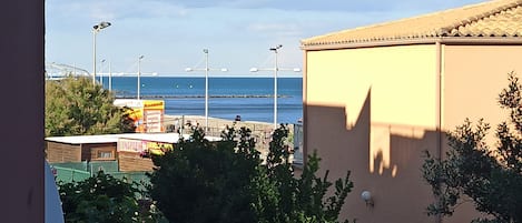 A view from a balcony overlooking a beach with a clear sky, a building with a tiled roof, and some greenery.