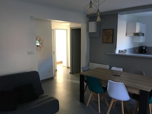 A modern living room with a sofa, a dining table with chairs, a kitchen area with cabinets and a coffee machine, and a hallway leading to an open door. - Logement Entier, cap D'agde, 6 Pers, 100m Plages, Falaises (Agde)
