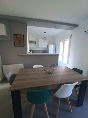 A modern dining area with a wooden table, white and green chairs, a kitchen in the background, and a ceiling light fixture. - Logement Entier, cap D'agde, 6 Pers, 100m Plages, Falaises (Agde)