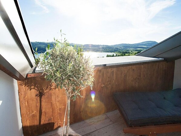 Une terrasse en bois offrant une vue sur un lac et des collines, une petite plante en pot et un coin salon avec des coussins.