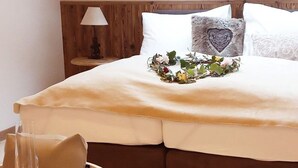 A bed with a beige bedspread, a heart-shaped pillow, and a floral arrangement. There is a bedside table with a lamp and a glass of water.