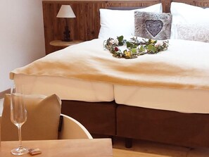 Un lit recouvert d’une couvre-lit beige, un oreiller en forme de cœur et un arrangement floral. Une table de chevet avec une lampe et un verre d’eau se trouve à côté du lit.