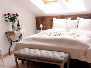 Een slaapkamer met een bed, een bankje, een nachtkastje, een lamp en een vaas met bloemen.
