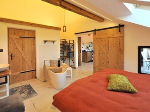 A bedroom with a bed, a red blanket, a wooden door, a white sink, and a kitchen in the background.