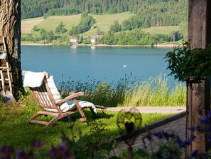 A wooden chair with a white cushion by a lake, surrounded by greenery and a tree. - Zur Linde - Roiderhof am Irrsee (Laiter)