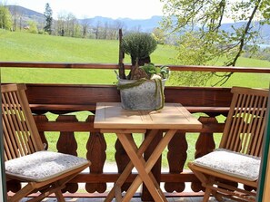 Una terraza de madera con dos sillas y una mesa, una planta en maceta y vistas a un campo verde y montañas.