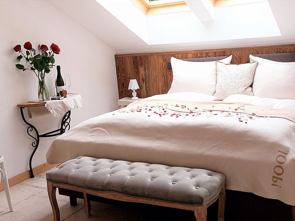 Un dormitorio con una cama, un banco, una mesita de noche, una lámpara y un florero con flores.