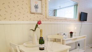 A white dining table with a bottle of champagne, a glass, and a vase with a red flower. A white chair with a red cushion is beside the table. A small white console table with a radio and a candle holder is against the wall. - Apartment Süd Terasse 45m² (Rømø)