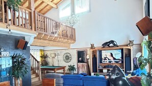 A spacious living area with a high wooden ceiling, a blue sofa, wooden dining table with chairs, and a staircase leading to an upper level. - Large atypical house in the Basque country, sleeps 16. Indoor pool (Ayherre)