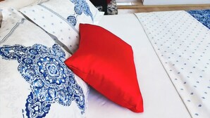 A bedroom with a bed, a nightstand, a wooden headboard, a red pillow, a white and blue patterned bedspread, a window with red curtains, and a framed picture on the wall. - Umut Denizi Konukevi (Bozcaada)