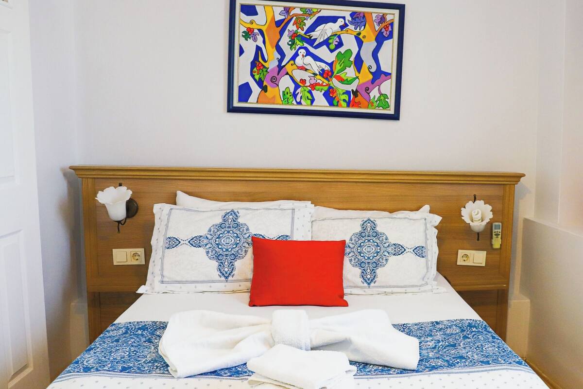 A neatly made bed with a blue and white patterned bedspread, a red pillow, and a decorative pillow with a blue design.