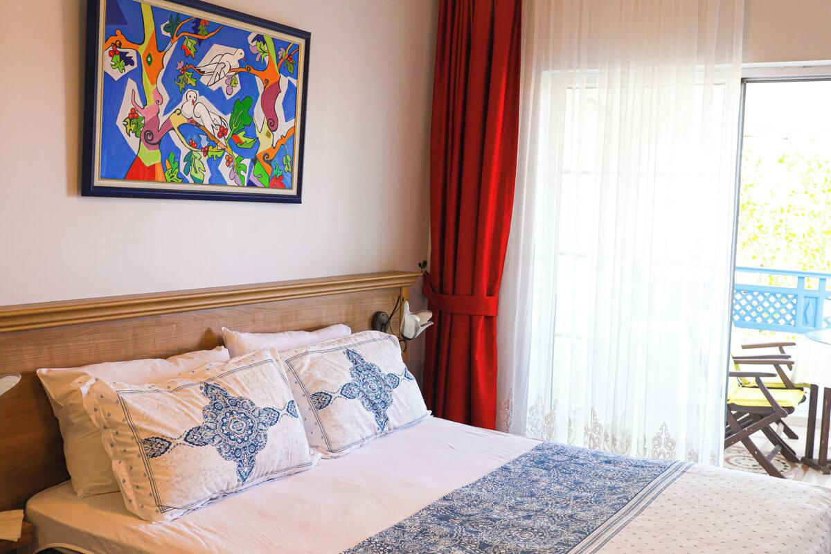 A hotel room with a bed, a painting on the wall, a window with red curtains, and a view of the outside.