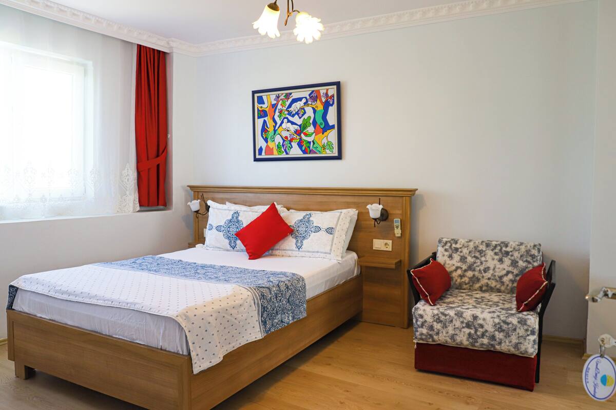 A bedroom with a wooden bed, a chair, a window with curtains, and a colorful abstract painting on the wall.