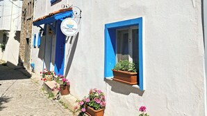 A building with blue window frames, a sign with a sun logo, and potted plants on the ground. - Umut Denizi Konukevi (Bozcaada)
