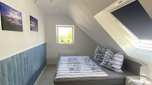 Une chambre avec un plafond en pente, un lit avec une literie sur le thème de la marine, une fenêtre donnant sur un paysage verdoyant et un ventilateur de plafond.