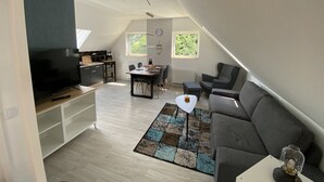 Un appartement moderne de style « loft » comprenant une cuisine, un coin repas et un salon. Il est équipé d’une grande télévision, d’une table à manger avec des chaises, d’un canapé et d’un tapis à motifs.