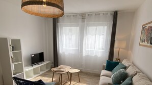 Ett vardagsrum med en soffa, en svart fåtölj, en tv, en lampa av rotting och ett fönster med tunna, genomskinliga gardiner.