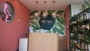 A reception area with a neon sign, a wall with a tropical leaf mural, and a staircase with potted plants. - Liberta Malioboro South (Yogyakarta)