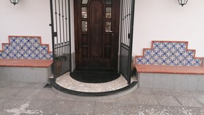Una puerta de madera con un panel de vidrio, flanqueada por dos lámparas tipo farol en color negro, que da paso a un vestíbulo con suelo de piedra y baldosas.