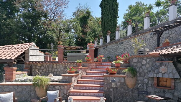 Una escalera de piedra con escalones de terracota que conduce a una zona cubierta, rodeada de plantas en macetas y mobiliario exterior.