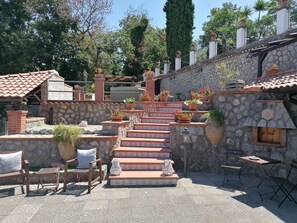Un escalier en pierre avec des marches en terre cuite menant à un espace abrité, entouré de plantes en pot et de mobilier d’extérieur.